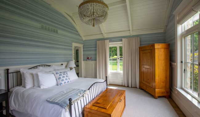 A bright bedroom with a vaulted white ceiling, blue-striped wallpaper, a black metal bed with white bedding, a wooden wardrobe, a matching chest, and large windows with beige curtains overlooking greenery.