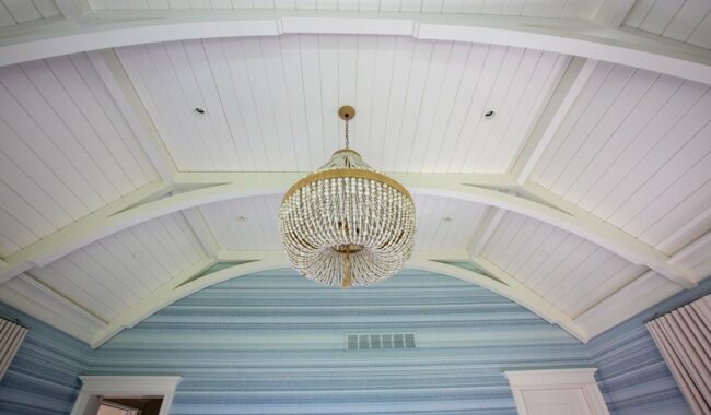 A large crystal chandelier hangs from a vaulted, white wooden ceiling above a room with blue striped wallpaper and white trim. Curtains frame two windows on either side of the image.