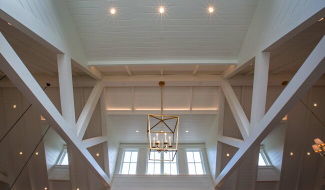 View of a high, vaulted white ceiling with exposed beams, a modern gold chandelier, and large windows letting in natural light, along with several recessed lights.