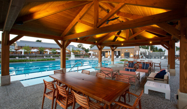 A covered patio with wooden tables and chairs overlooks a large outdoor swimming pool. The patio features warm ceiling lights, and loungers line the side of the pool. Buildings are visible in the background.