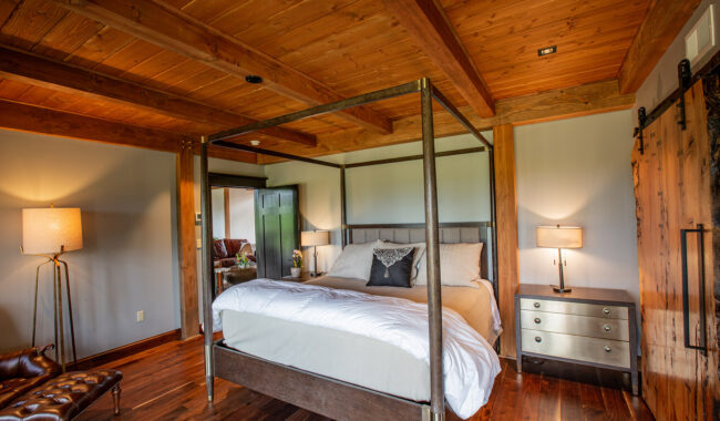 A cozy bedroom with wooden floors and ceiling, featuring a four-poster bed with white bedding, two nightstands with lamps, a leather chair, and a sliding barn door. Natural light fills the warm, rustic space.