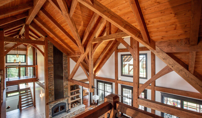 A spacious interior of a modern rustic home with exposed wooden beams, high vaulted ceiling, large windows, cozy stone fireplace, and a loft area overlooking the living space. Natural light fills the room.