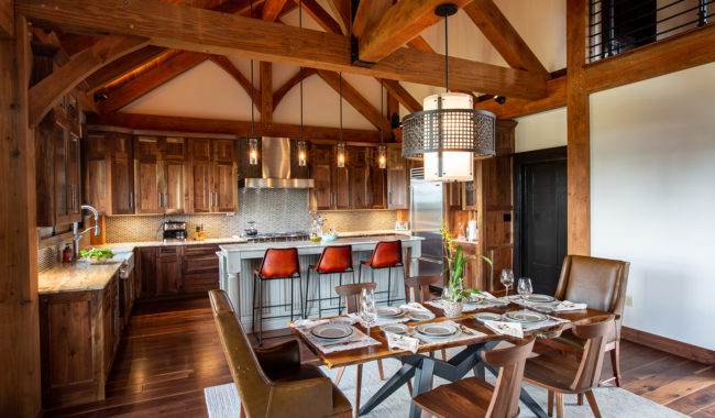 A rustic kitchen and dining area with exposed wooden beams, brown cabinets, a kitchen island with three orange stools, a dining table set for six, and a modern chandelier above. Natural light fills the cozy space.
