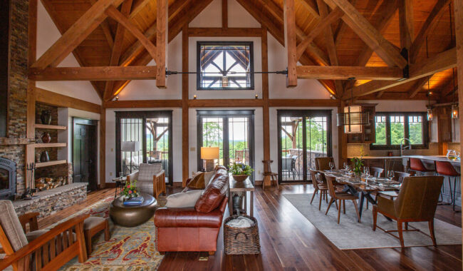 Spacious rustic living and dining room with exposed wooden beams, brown leather sofa, armchairs, stone fireplace, large windows, and a dining table set for a meal; kitchen area visible in the background.