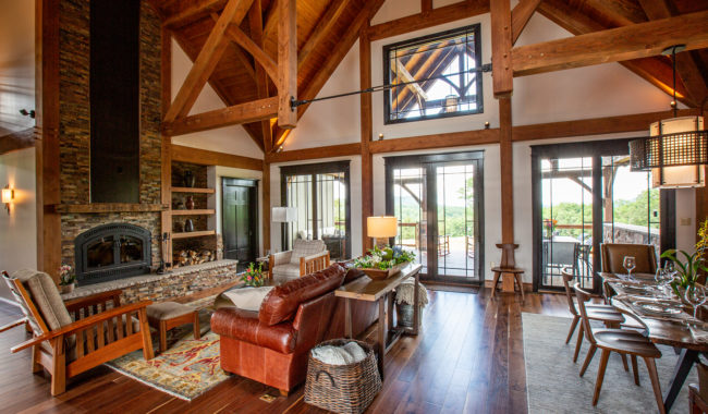 Spacious living room with high wooden ceilings, exposed beams, large windows, stone fireplace, leather and fabric seating, dining area set for a meal, and hardwood floors. Natural light fills the cozy, rustic interior.