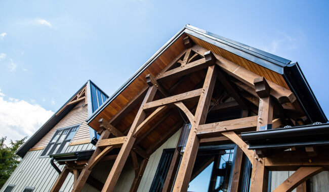 Close-up view of a modern timber-frame house with exposed wooden beams, metal roof, large windows, and clear blue sky in the background.