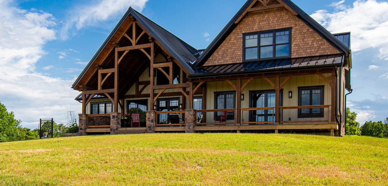 A modern rustic house with wooden beams and a large covered porch sits on a grassy hill under a blue sky with scattered clouds.