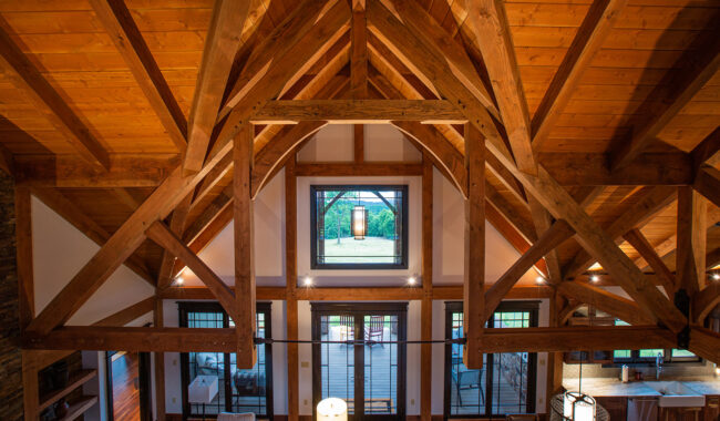 A warm, rustic living space with exposed wooden beams, a high vaulted ceiling, large windows, and glass doors opening to a patio. Natural light fills the room, highlighting wooden interiors and modern furnishings.
