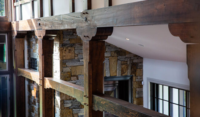 A close-up view of a rustic interior featuring exposed dark wooden beams and columns, with a stone wall and large windows, creating a warm and inviting atmosphere.