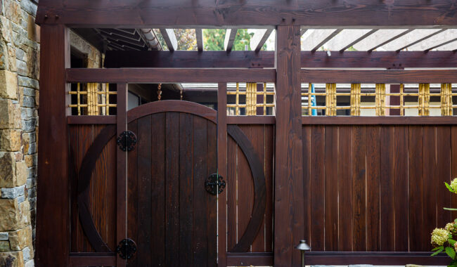 A wooden garden gate with a circular design, surrounded by a tall wooden fence with rectangular lattice accents on top. Stone pillars and a pergola with translucent panels are also visible.