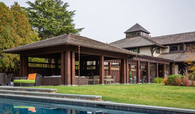 A spacious backyard features a wooden pergola with outdoor seating and dining areas, adjacent to a swimming pool, surrounded by grass and trees, next to a large house.