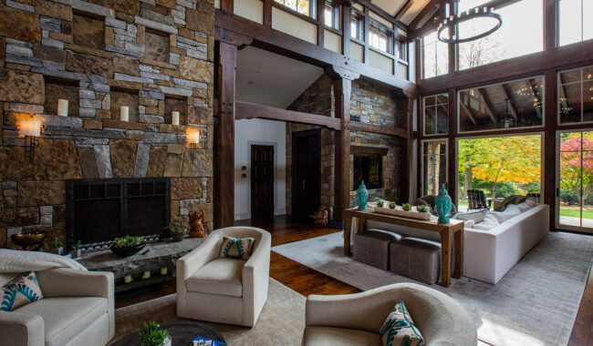 Spacious, modern living room with high ceilings, large stone fireplace, tall windows, comfortable beige seating, a long dining table, and abundant natural light. Greenery visible outside enhances the warm, inviting atmosphere.