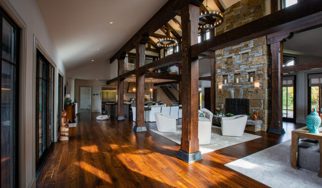 Spacious open-concept living area with exposed wooden beams, a stone fireplace, modern white furniture, hardwood floors, large windows, and a vaulted ceiling, blending rustic and contemporary design elements.