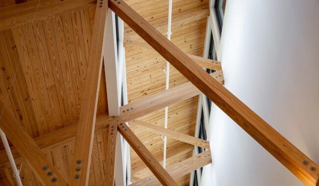 Exposed wooden beams and trusses form a geometric pattern on the ceiling, with natural wood grain visible. Metal bolts connect the beams. White pipes and part of a white wall are also seen.