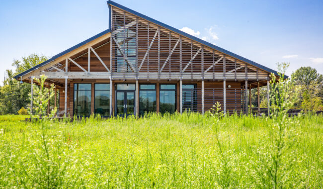 A modern wooden building with large windows and a distinctive sloped roof stands behind a lush green field, under a clear blue sky.