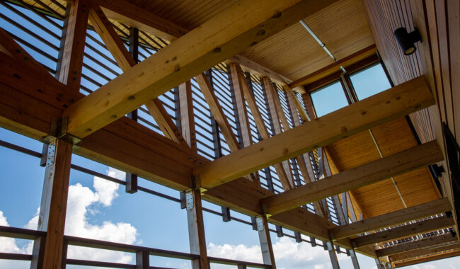 Wooden beams and slatted panels form the roof and walls of a modern structure, letting sunlight through. Blue sky and white clouds are visible through the open sections of the building.