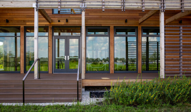 A modern building with large glass doors and windows, wooden siding, a raised deck with steps, and green plants in front. The glass reflects a grassy landscape and blue sky with clouds.