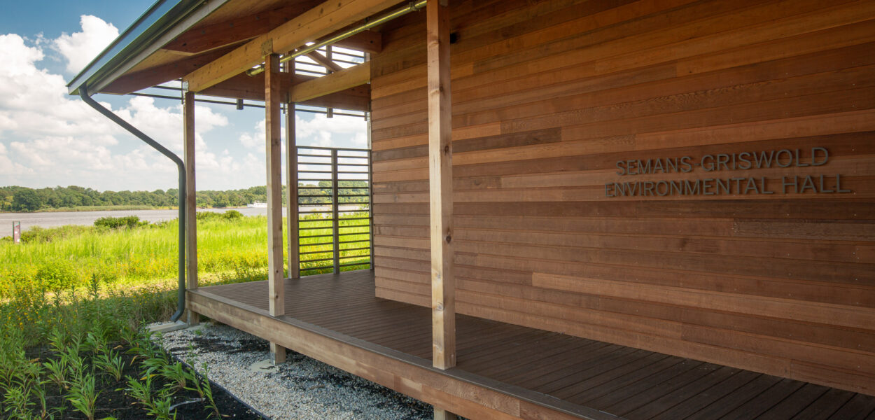 A modern wooden building labeled “Semans-Griswold Environmental Hall” stands amid green grass and plants, with a partly cloudy sky and open field in the background.