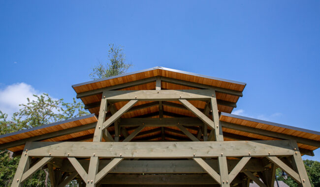 A wooden gazebo with a layered roof is shown against a clear blue sky, surrounded by green trees and part of a house visible on the right side in the background.