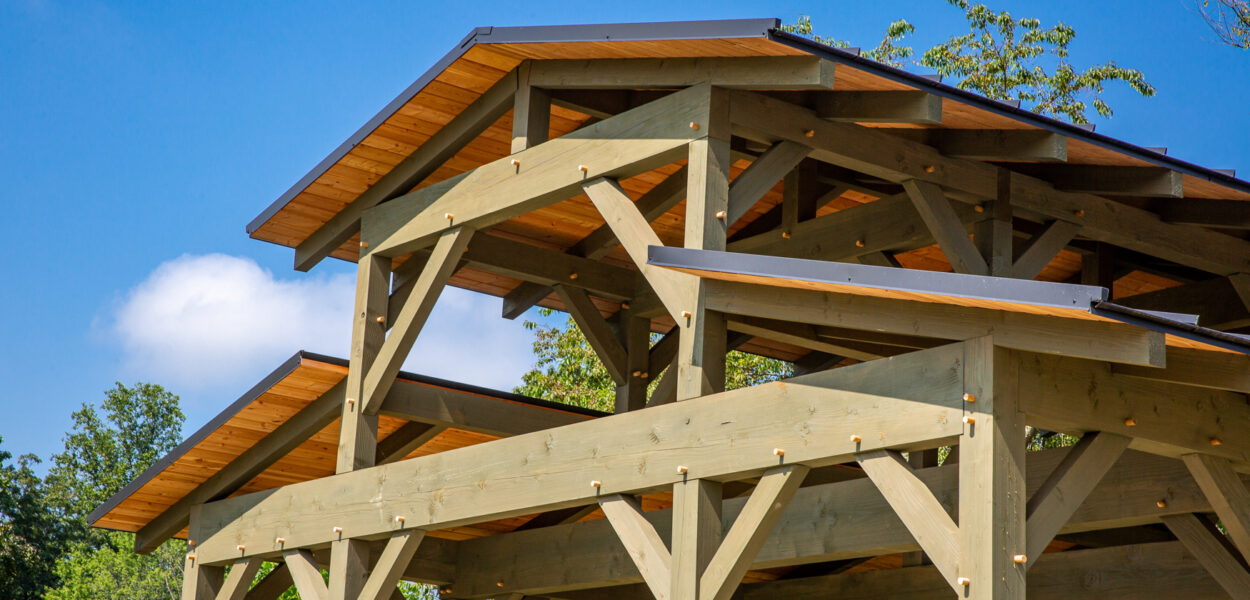 A close-up of a wooden pavilion structure with exposed beams, multiple gabled roofs, and a clear blue sky in the background. Trees and greenery are visible behind the structure.