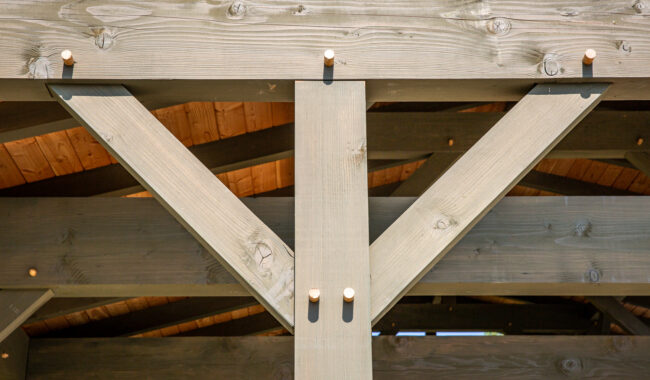 Close-up of a wooden pergola structure, showing intersecting beams joined with wooden pegs. Sunlight highlights the wood grain and craftsmanship, with a roof and sky partially visible in the background.