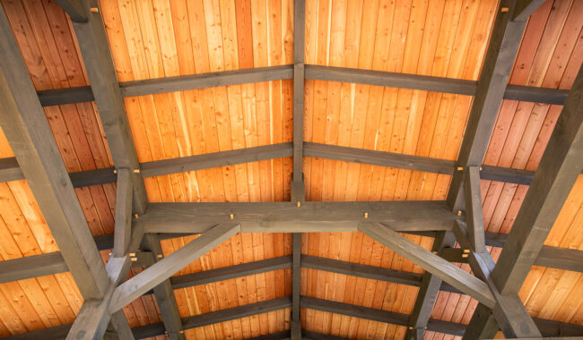 View looking up at a wooden ceiling with exposed dark beams and natural wood panels, forming a symmetrical, peaked roof structure.