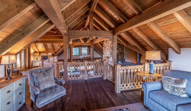 A cozy loft with exposed wooden beams, rustic railings, a stone fireplace, armchairs, lamps, and warm lighting, overlooking a spacious living area below.