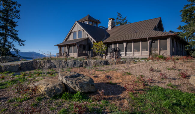 A rustic house with a metal roof sits on a rocky, landscaped hillside under a clear blue sky, surrounded by trees and distant mountains.
