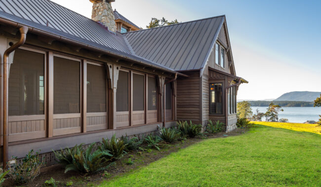 A brown wooden house with a metal roof sits on a grassy lawn near a body of water, surrounded by trees and mountains under a clear blue sky.