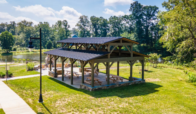 A wooden pavilion with picnic tables underneath sits on a grassy area near a pond, surrounded by trees on a sunny day. A sidewalk and a black lamp post are visible in the foreground.