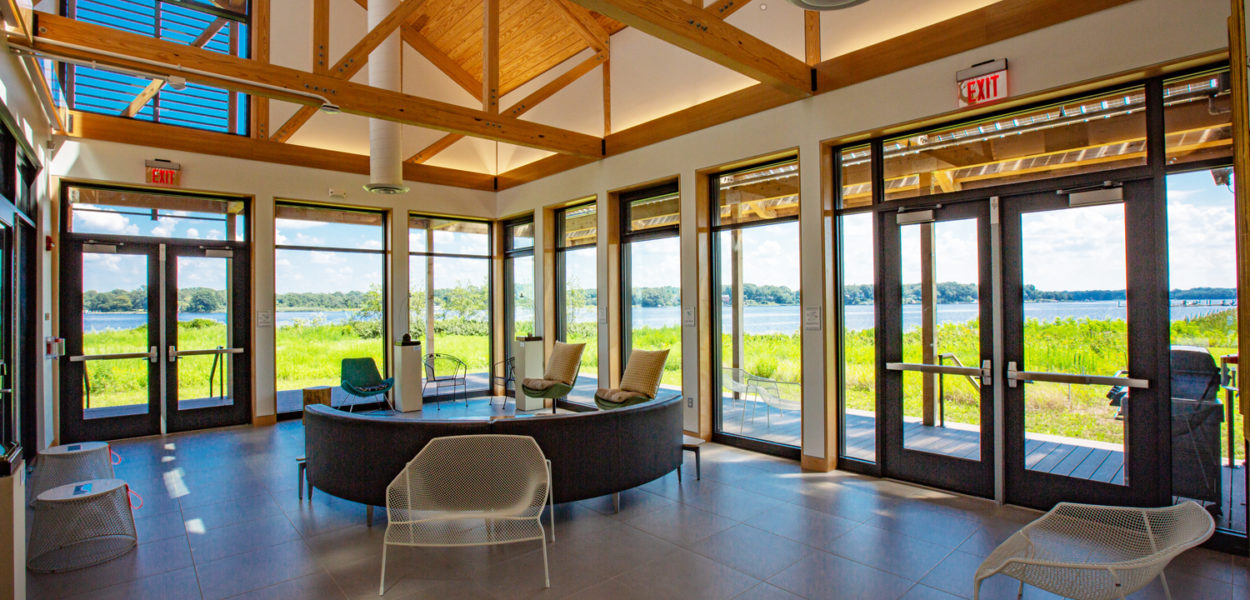 A bright, open room with large windows, wooden beams, and modern seating, offering scenic views of a lush green landscape and water outside under a blue sky.