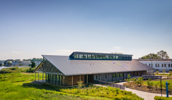 Modern, single-story building with a sloped metal roof and large windows, surrounded by green grass and shrubs, under a clear blue sky. A path leads to the entrance, and water and other buildings are visible in the background.