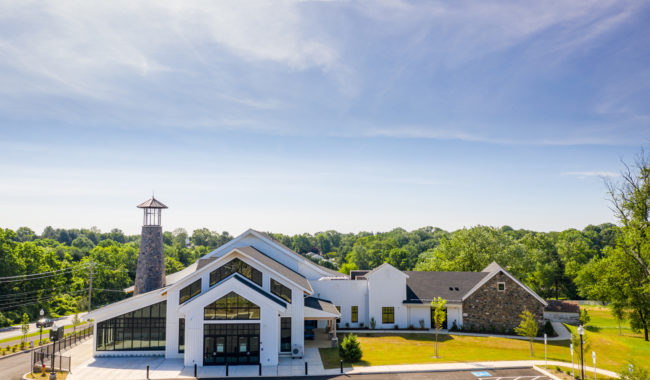 A modern church building with large glass windows and a stone tower, surrounded by trees under a blue sky, with an empty parking lot in the foreground.