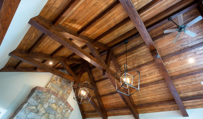 Exposed wooden beams and paneling on a vaulted ceiling with two geometric pendant lights and a ceiling fan; a stone fireplace rises along one wall.