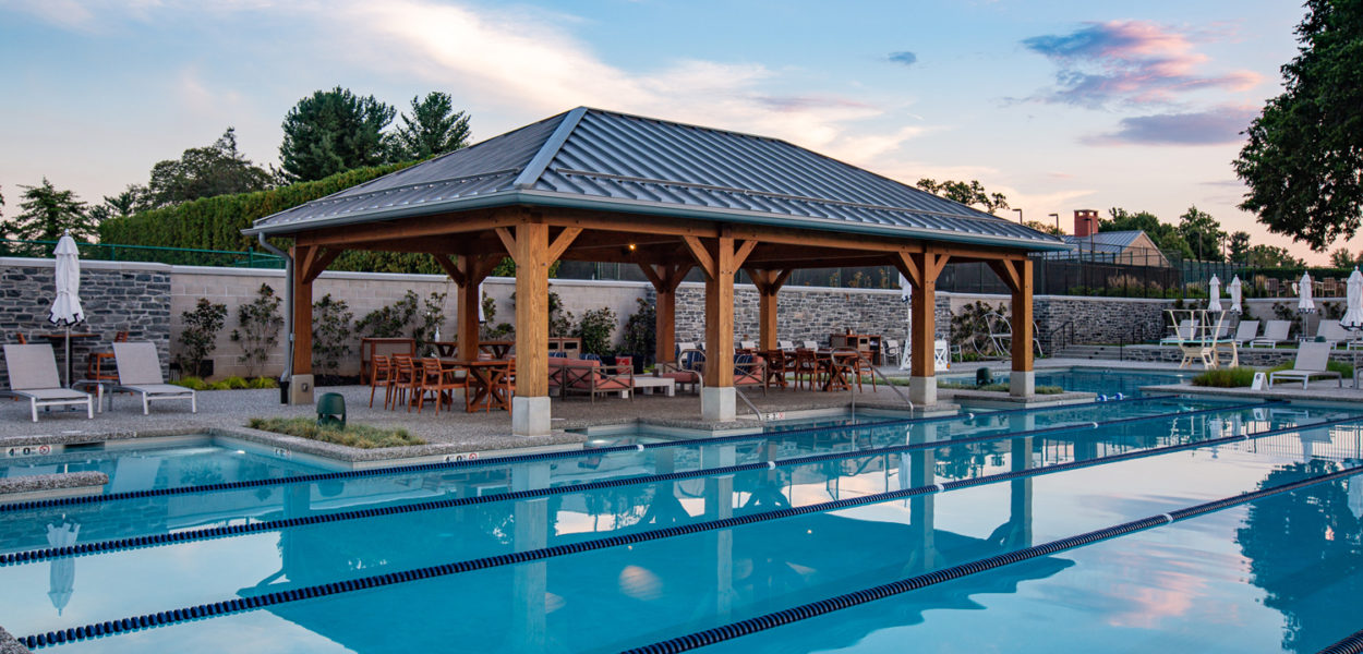 Outdoor swimming pool with swim lanes, lounge chairs, and a wooden pavilion with tables and chairs underneath. The scene is set at sunset, with trees and a stone wall in the background.