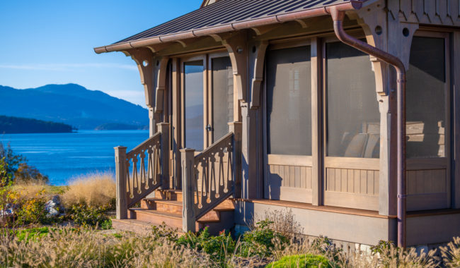 A wooden house with a screened porch overlooks a bright blue lake or sea, with mountains and clear sky in the background. Tall grasses and plants surround the house on a sunny day.