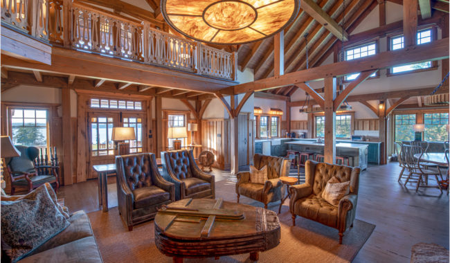 Rustic living room with high wooden ceilings, exposed beams, large circular chandelier, leather armchairs, a wooden coffee table, and an open kitchen and dining area in the background. Sunlight fills the room.