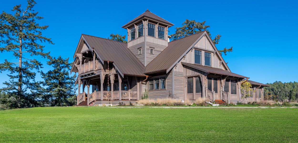 A large wooden house with a central tower stands on a well-manicured green lawn, surrounded by trees, under a clear blue sky.