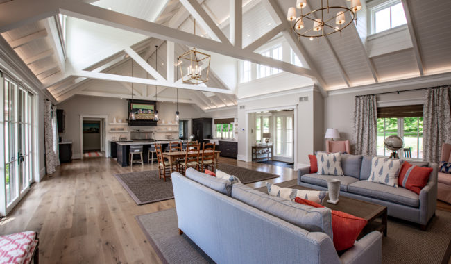 A spacious, bright living room with high vaulted ceilings, exposed beams, large windows, light wood floors, and a mix of blue, pink, and neutral furnishings. The space is open to a kitchen and dining area.