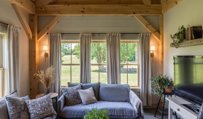 A cozy living room with wooden beams, large windows with light curtains, two gray sofas with patterned pillows, indoor plants, and a wall-mounted TV. Sunlight streams in, highlighting the greenery outside.