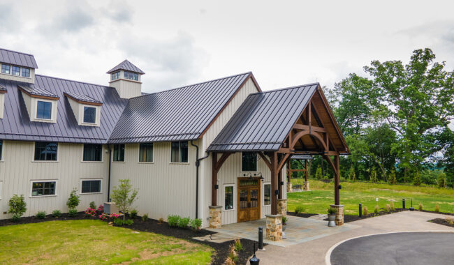 A modern barn-style building with metal roofing, large windows, and a covered entrance supported by wooden beams, surrounded by greenery and a curved driveway.