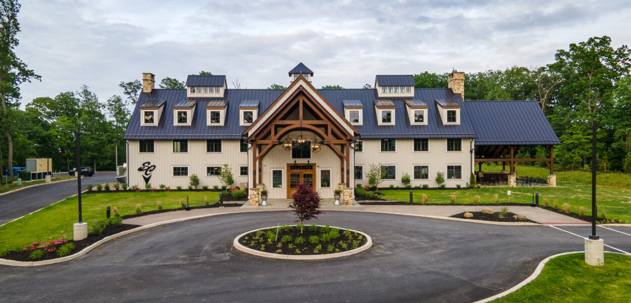 A large, modern barn-style building with a metal roof, white walls, and wooden beams stands at the end of a circular driveway, surrounded by green grass, trees, and landscaped flower beds.