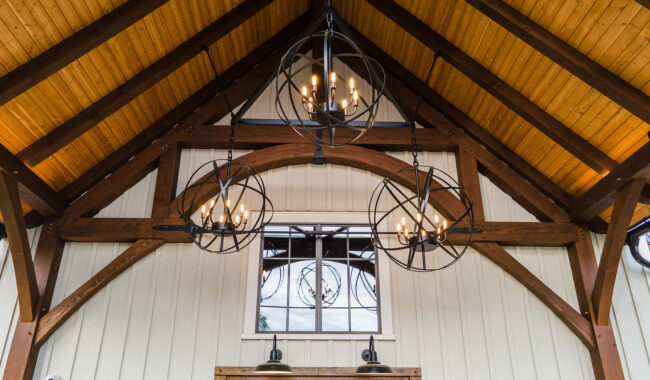 Wood-paneled vaulted ceiling with exposed beams, featuring three modern spherical chandeliers with candle-shaped bulbs, and a window letting in natural light above rustic wall sconces.