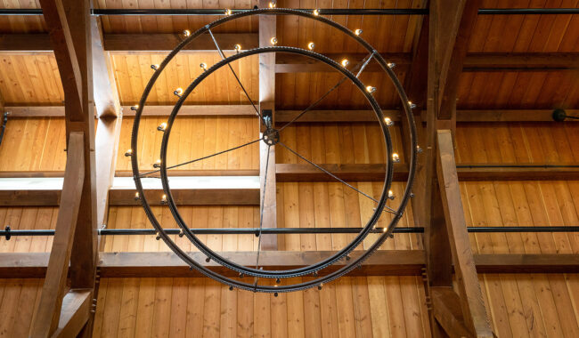 A large, circular metal chandelier with small exposed bulbs hangs from a wooden ceiling with visible beams. The warm-toned wood creates a rustic, inviting atmosphere.