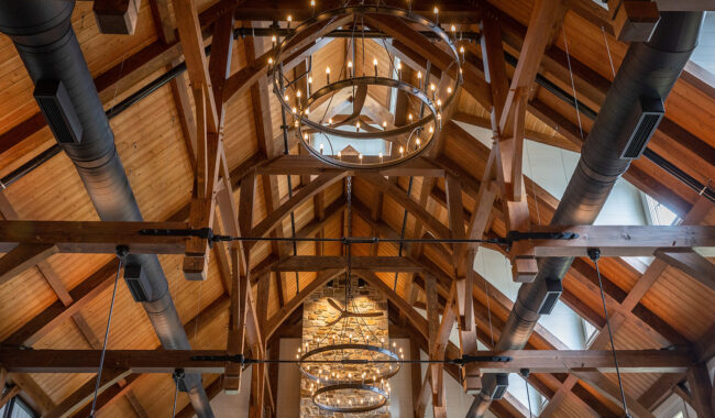 Vaulted wooden ceiling with exposed beams, black ducts, and large circular chandeliers with warm lights, above a stone fireplace wall with windows on the sides.