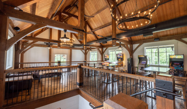 A spacious, rustic game room with exposed wooden beams, high ceilings, large windows, arcade machines, bar seating, and comfortable lounge furniture, illuminated by modern chandeliers.