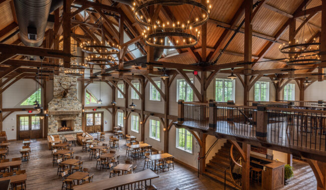 A spacious, rustic dining hall with wooden beams, large chandeliers, a stone fireplace, rows of wooden tables and chairs, and a lofted seating area with stairs leading up. Natural light streams in through large windows.
