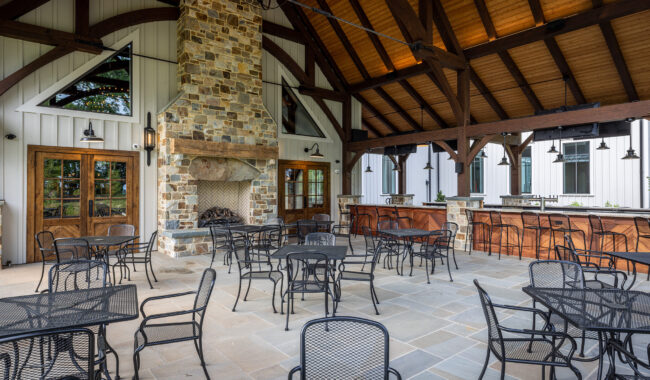 A covered outdoor patio with a high wooden ceiling, stone fireplace, and multiple black metal tables and chairs. Large windows and doors line the white-paneled building, creating an inviting dining or gathering space.