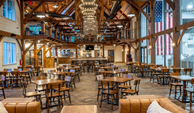 Spacious, rustic dining hall with wooden beams, high ceilings, chandeliers, and many wooden tables and chairs. A large American flag hangs on the right, and a mezzanine overlooks the main floor.