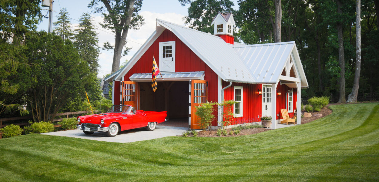 A bright red classic car is parked in front of a red barn-style garage with white trim and an open door, surrounded by neatly mowed grass and tall green trees.
